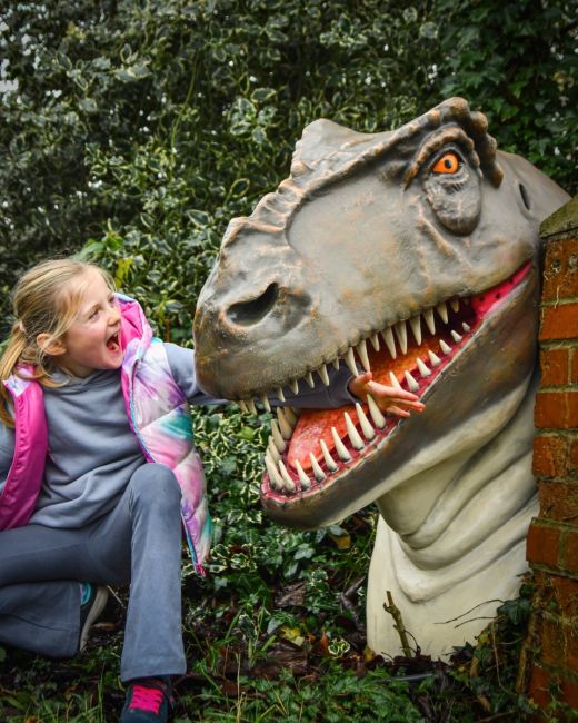 ‘Reginald” T-Rex Head Garden Wall Mount shows t-rex and child pretending to have their arm bitten