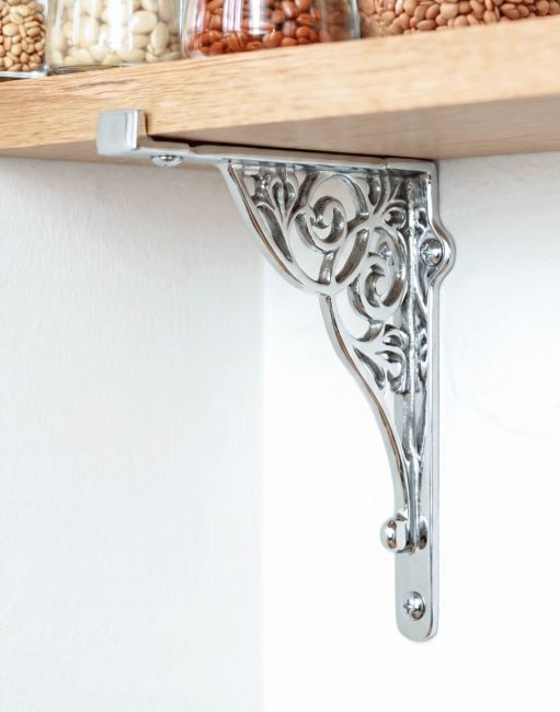 The 27cm x 22cm Entwined Scroll Bright Chrome Shelf Bracket Mounted on a White Coloured Kitchen Wall Supporting a Wooden Shelf - Digital Illustration