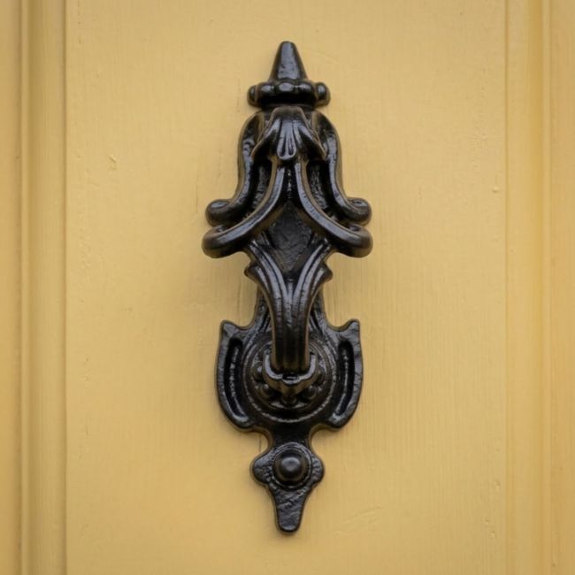 The Traditional Black Iron Gothic "Rawnsley Pattern" Door Knocker Shown Mounted onto a Yellow Door on the Front of a Home - Digital Illustration