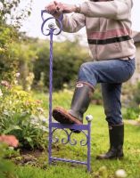 A Lilac Boot Scraper Shown Being Used by a Gardener Next to a Flower Bed - Digital Illustration