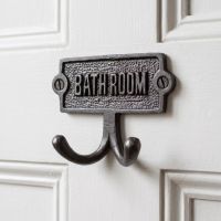 The Bathroom Plaque Double Robe Hook in Cast Antique Iron Shown Mounted onto the Back of a white Door - Digital Illustration