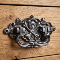 The Cast Antique Iron Skull & Crossbones Drop Handle - Antique Iron Finish Shown on a Wooden Drawer - Digital Illustration