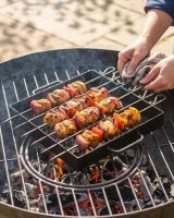 The Kadai Hand-Forged 6-Piece Skewer Set with Elevated Rack Shown Being Used above a Lit Fire Bowl with Hands in Shot for Scale - Digital Illustration