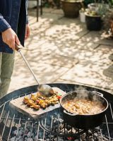 The Kadai Long-Handled Iron Cooking Ladle with Wooden Grip in Front of a Garden Patio Being Uded to Serve a Stew - Digital Illustration