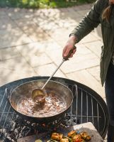 The Kadai Long-Handled Iron Cooking Ladle with Wooden Grip Shown Being Used on a Sunny Patio - Digital Illustration