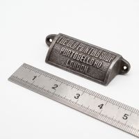 The Portobello Antique Iron Cup Pull Handle 98mm on a White Background next to a Steel Ruler for Scale - Digital Illustration