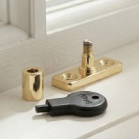 The "Abberwick" Locking Stay Pin Polished Brass Plced in Front of a White Window Frame in a Country Home - Digital Illustration