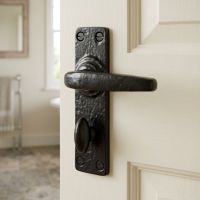 The "Castello" Smooth Bathroom Lever Lock Set Door Handle on a Cream Wooden Door With a Magnolia Bathroom Behind - Digital Illustration