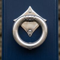 The "Lorien" Ring Door Knocker - Polished Stainless Steel on a Dark Blue Front Door on a Sunny Day - Digital Illustration 
