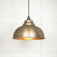 The Arcus Spun Pendant Light in Aged Brass Shown Lit up on a White Panelled Background