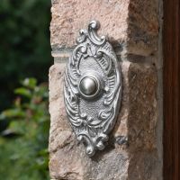 The Clarice Oval Satin Chrome Ornate Bell Push Fitte to a Sunny Stone Wall next to a Front Door - Digital Illustration