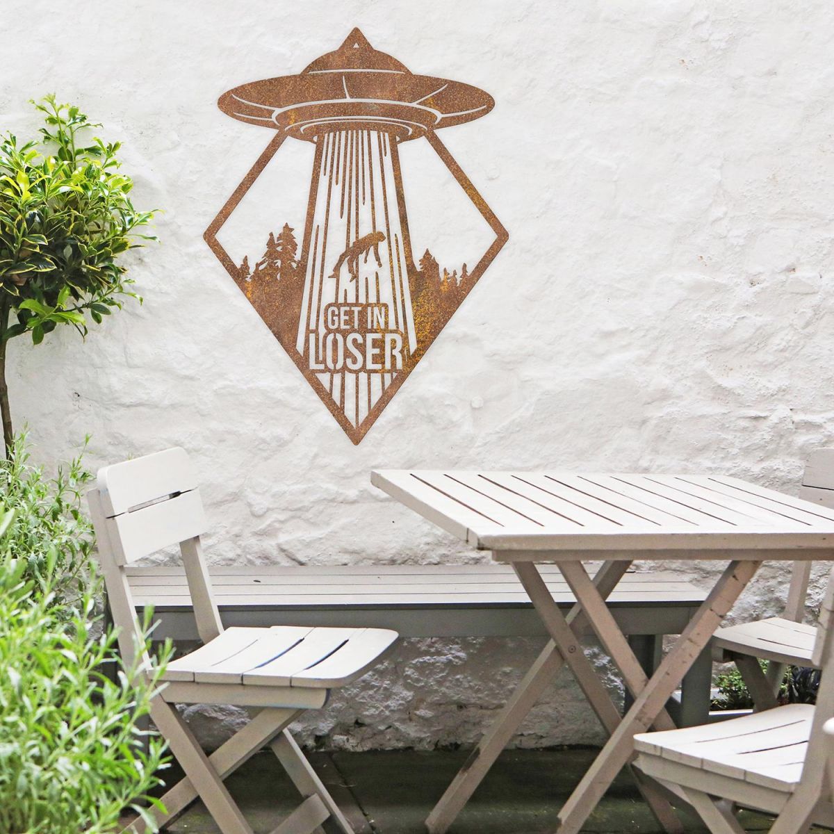 "Get in Loser" Alien Wall Art in Situ by a Wooden Table Set in the Garden