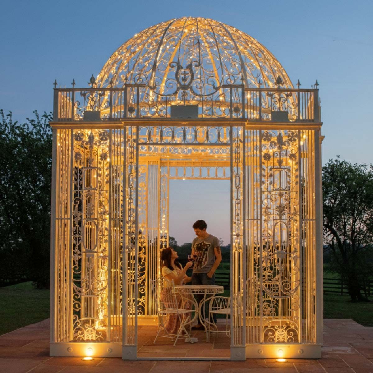 Digital representation of "The Royal Alexandria" Cream Wrought Iron Gazebo lit up with fairy lights at night