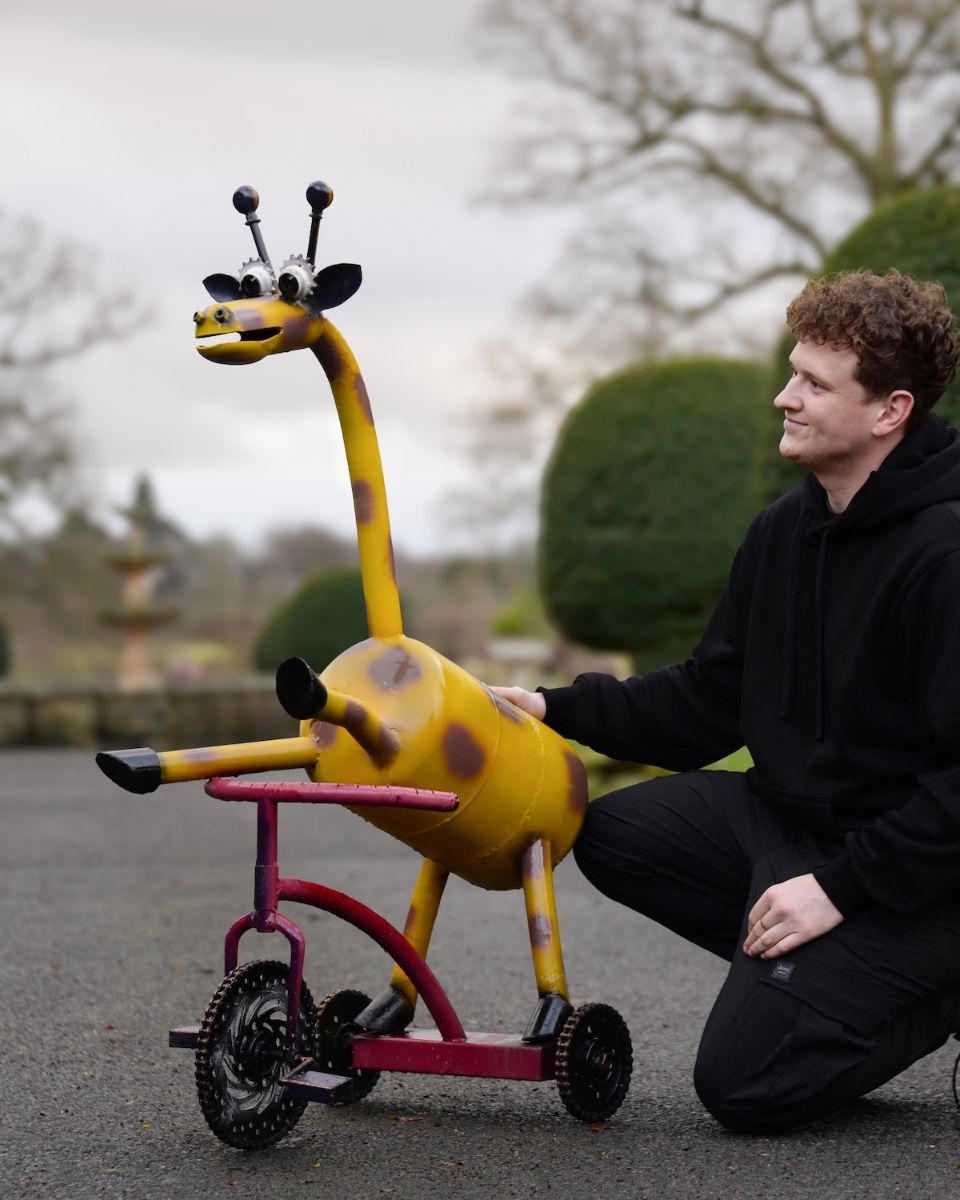 ‘Freewheeler” the Upcycled Metal Giraffe Garden Sculpture on a Tricycle to scale with man crouching next to sculpture for perspective
