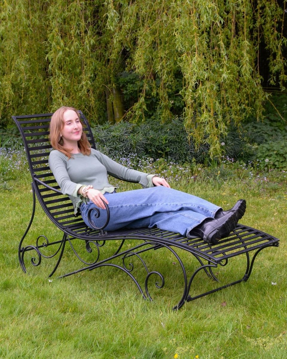 The "Windsor" Scrollwork Wrought Iron Sun Lounger in a Black Finish Positioned on a Sunny Garden Lawn with a Lady Relaxing on it 