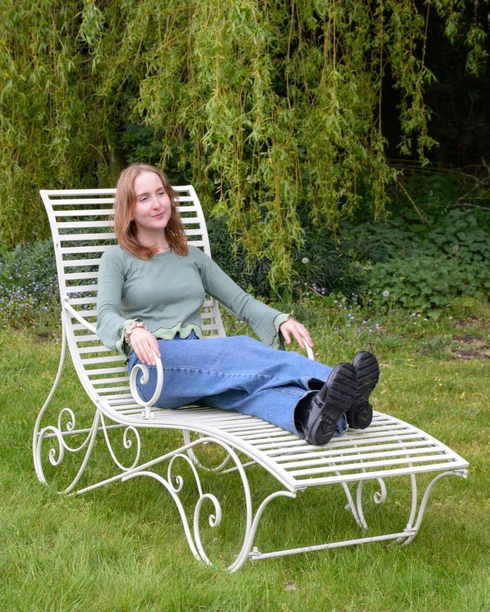 The "Windsor" Scrollwork Wrought Iron Sun Lounger in a Cream Finish Positioned on a Sunny Garden Lawn with a Lady Relaxing on it