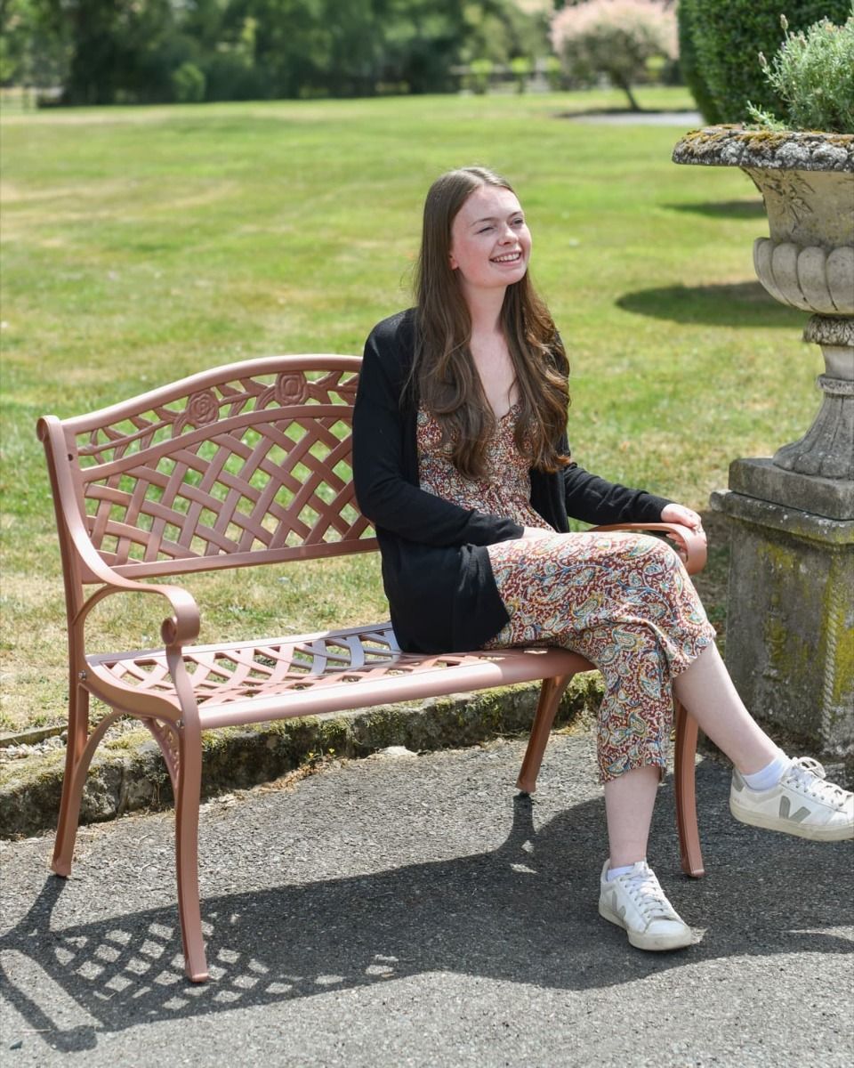 The Antique Pink "Thorpe" Two Seater Garden Bench Shown with a Smiling Lady Sat on it in the Sun