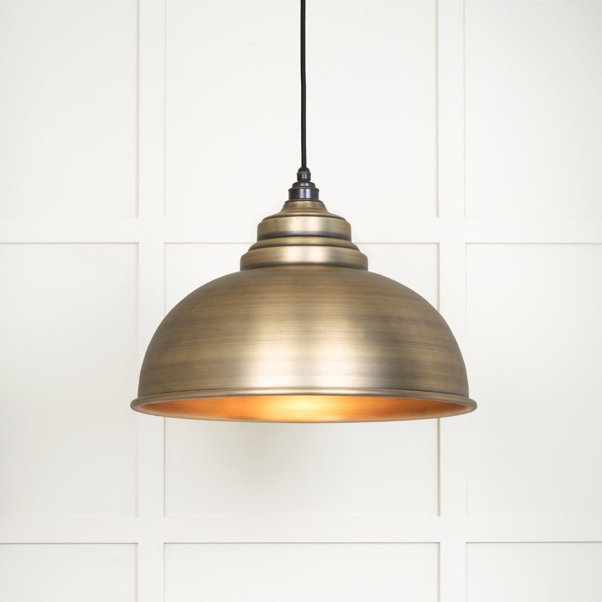 The Arcus Spun Pendant Light in Aged Brass Shown Lit up on a White Panelled Background
