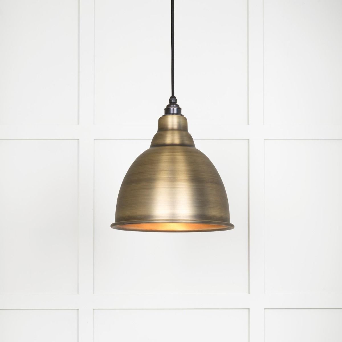 The Aura Dome Pendant Light in Aged Brass Shown Lit up on a White Panelled Background