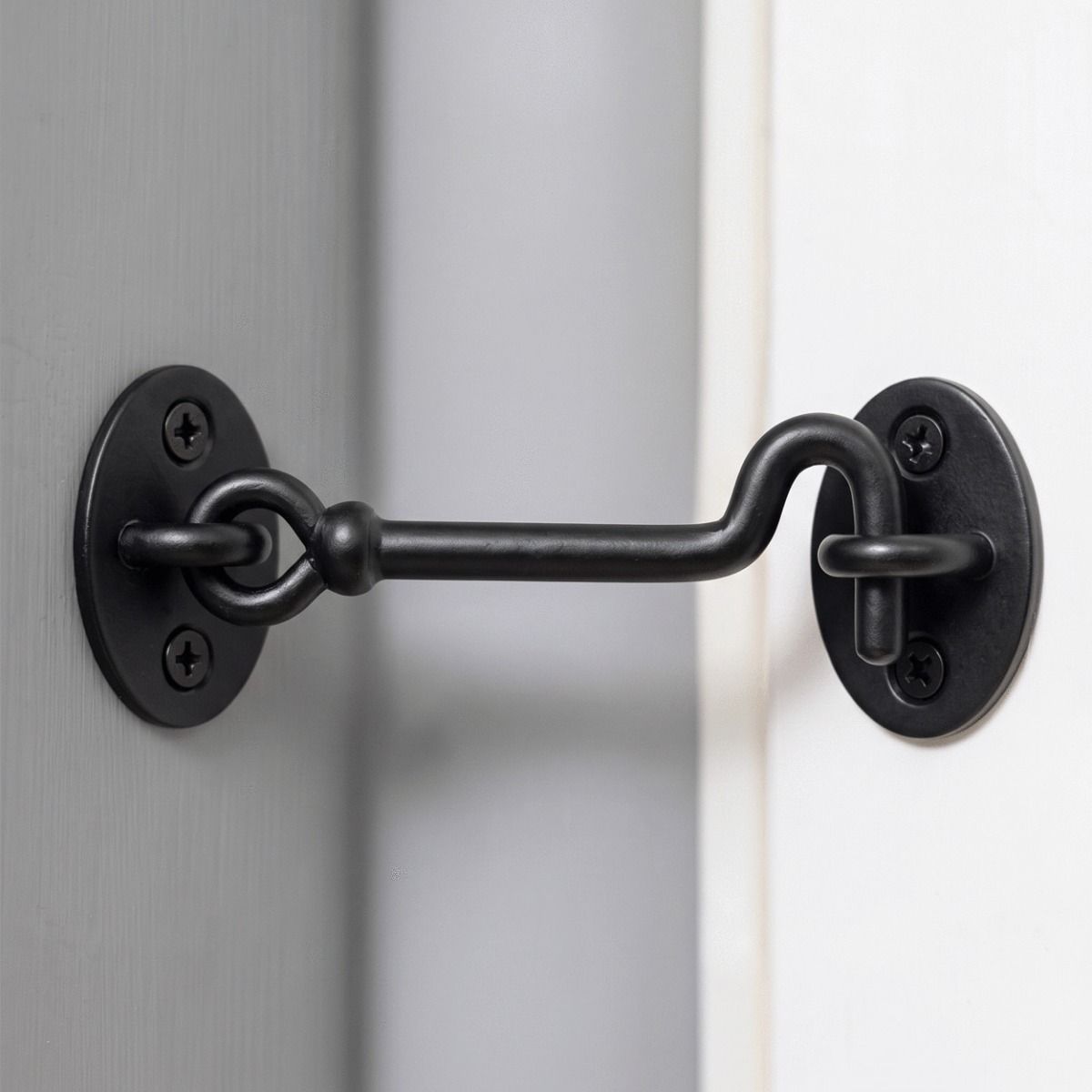 The Matte Black Cabin Hook and Eye Latch Shown Fitted in a Modern Home on a Grey Wall next to a White Door - Digital Illustration 