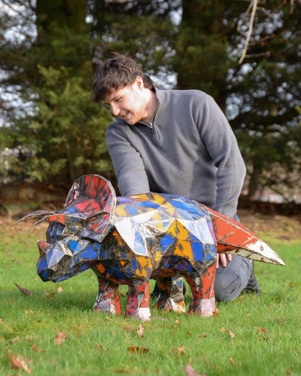 "Trike’ the Triceratops Upcycled Oil Drum Garden Sculpture with man crouching down next to sculpture for scale